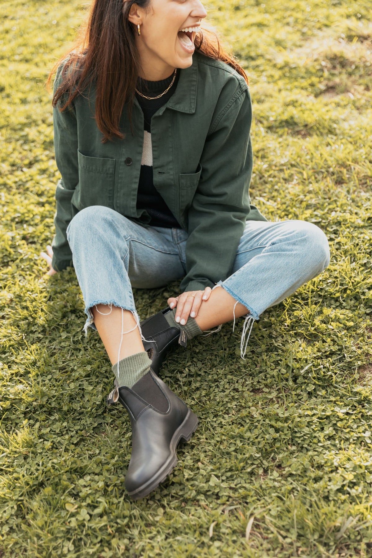Woman sitting on grass wearing black Blundstone Chelsea boots with light-wash jeans and olive socks