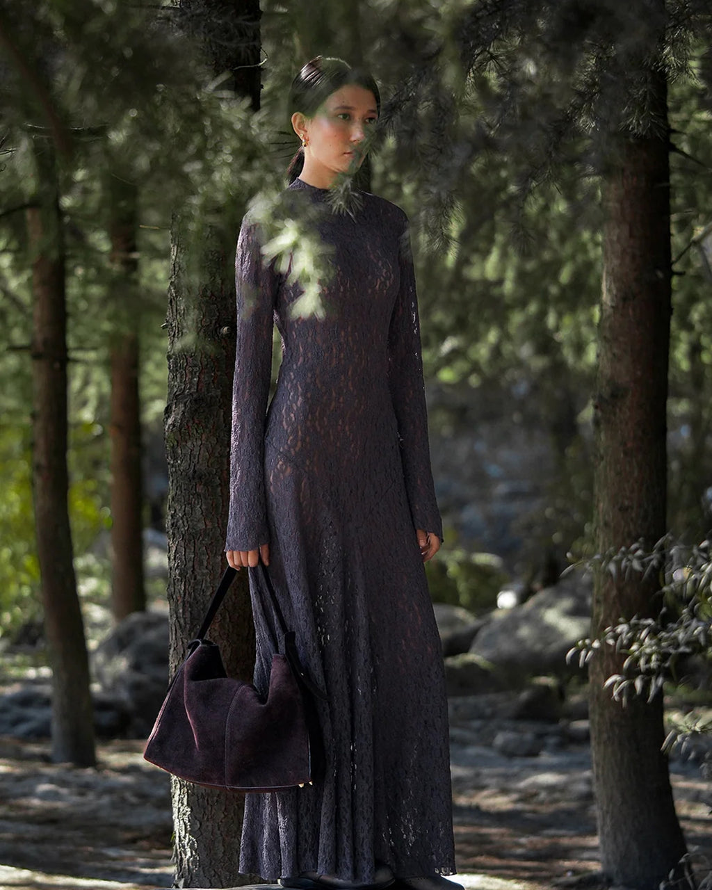 Model holding the ALEO Shiloh Suede Shoulder Bag in Crema by its handle in a pine forest