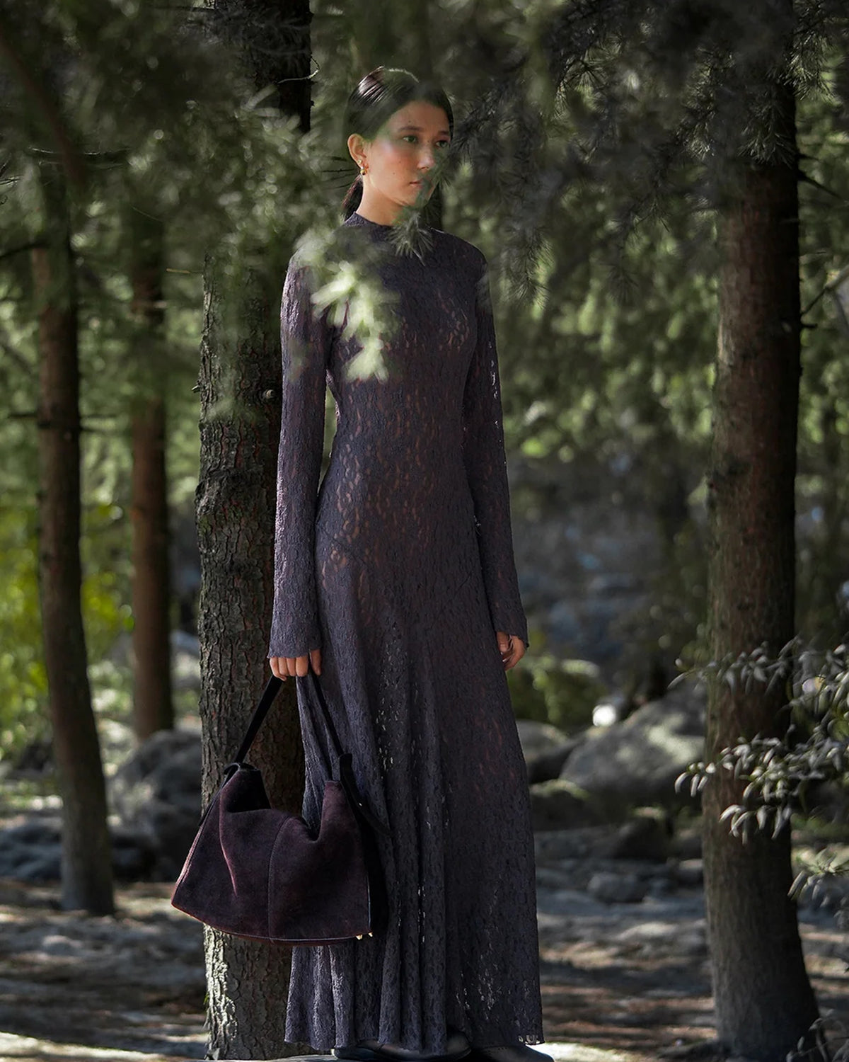 Model holding the ALEO Shiloh Suede Shoulder Bag in Crema by its handle in a pine forest