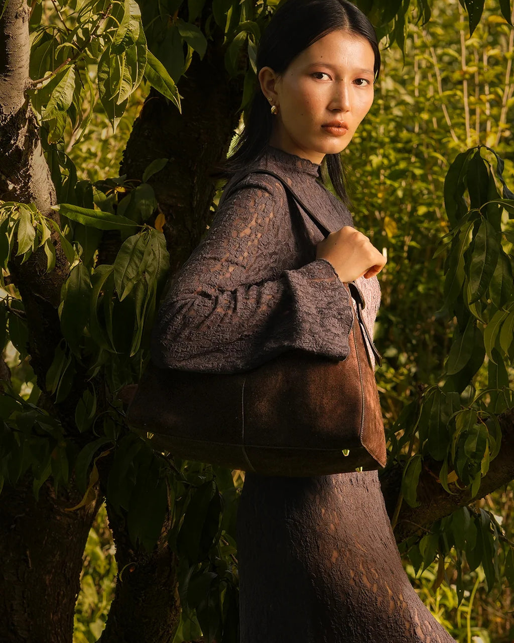 Model carrying the ALEO Shiloh Suede Shoulder Bag in Crema on her shoulder among leafy branches