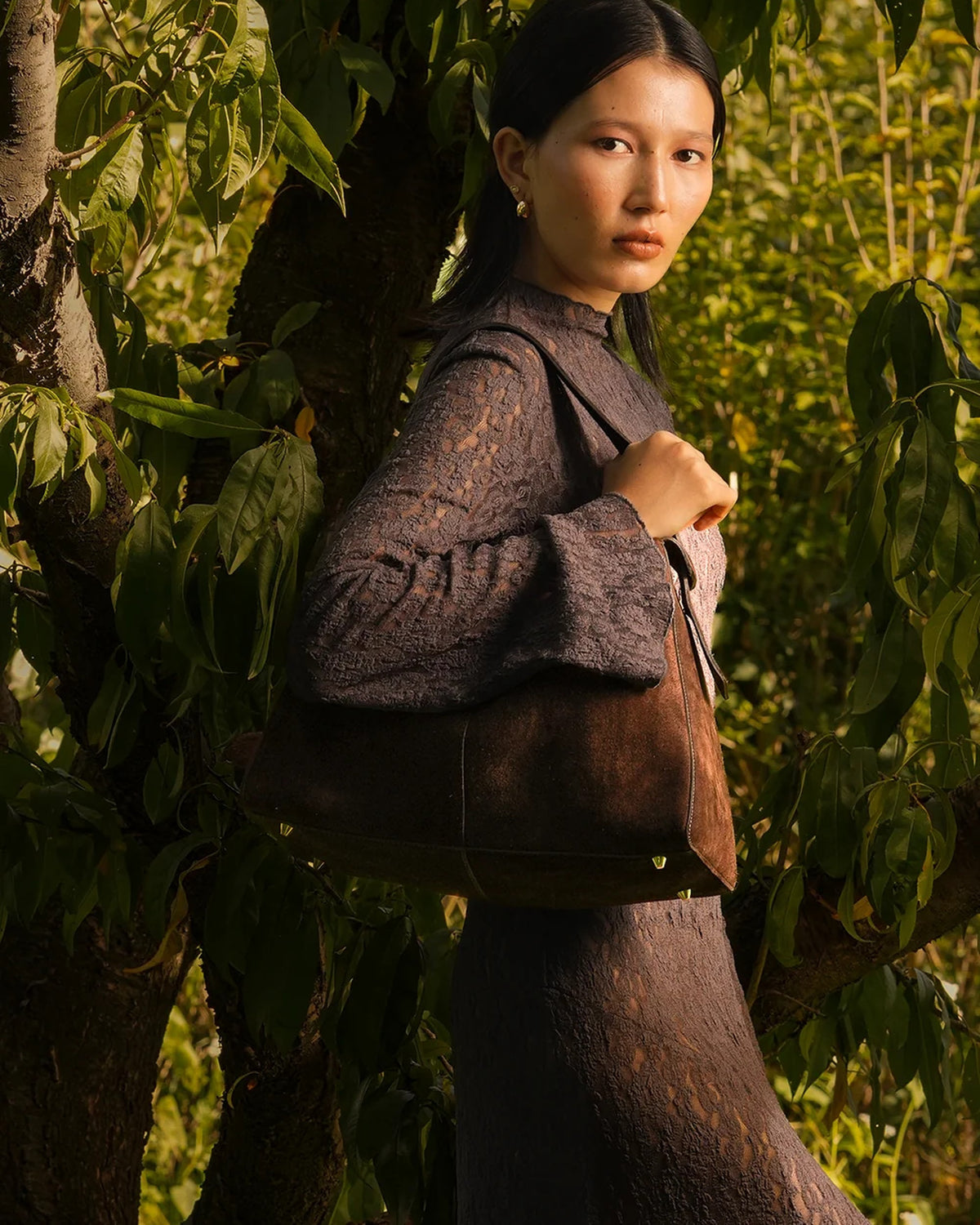 Model carrying the ALEO Shiloh Suede Shoulder Bag in Crema on her shoulder among leafy branches