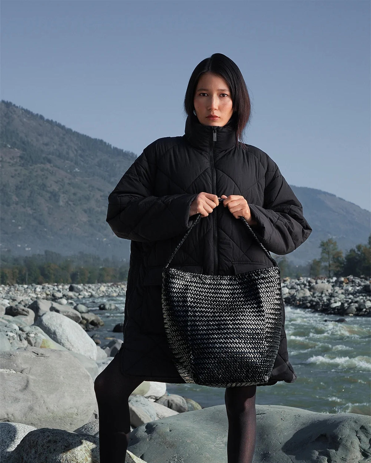 Model holding the ALEO Toka Woven Tote in Black by its strap in front of her, river and mountains behind