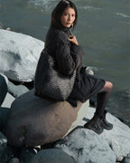 Model seated on a boulder by a river carrying the ALEO Toka Woven Tote in Black on her shoulder