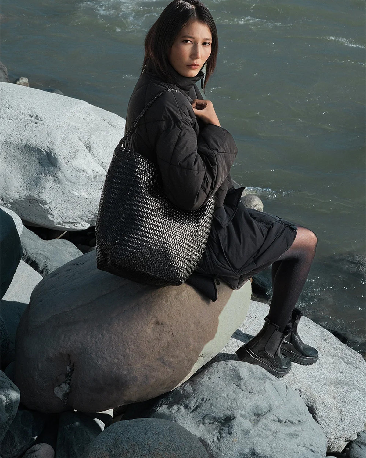 Model seated on a boulder by a river carrying the ALEO Toka Woven Tote in Black on her shoulder