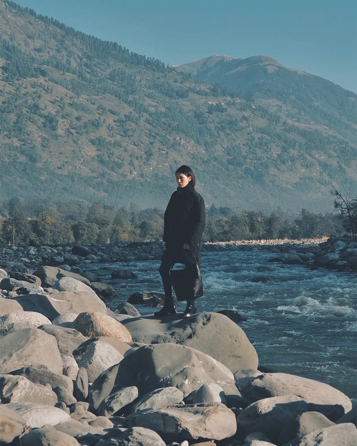 Model standing on rocks by a river holding the ALEO Toka Woven Tote in Black