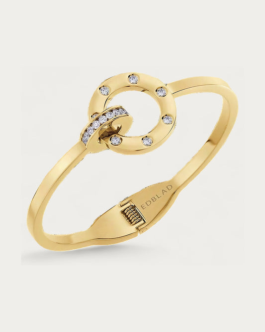 Edblad Ida Bangle Bracelet In Gold, a rigid gold bangle with connected rings set with clear stones, product photo on a soft off-white background