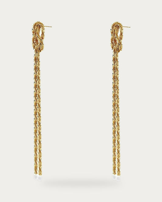 Edblad Tinsel Festive Earrings In Gold, a pair of gold earrings with a small knot and a long tinsel chain, product photo on a soft off-white background